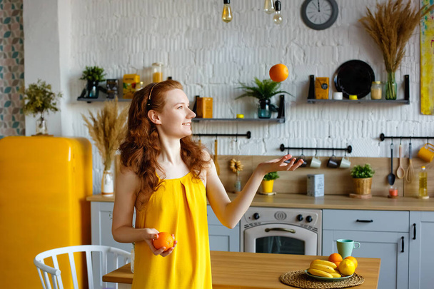 Portrait d'une jolie fille aux cheveux roux en bonne santé dans une robe de soleil jaune avec des fruits dans les mains dans la cuisine. Jette une orange. - Photo, image