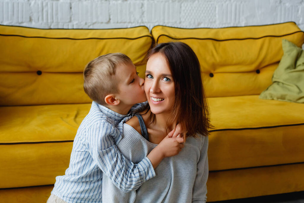Mutter und Sohn zusammen auf dem Boden neben dem gelben Sofa. Der Junge küsst seine geliebte Mutter auf die Wange. - Foto, Bild