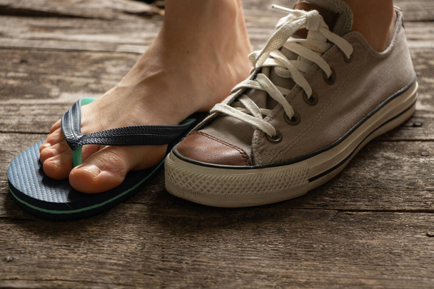 jedna damska stopa w brązowych tenisówkach tekstylnych, a druga w klapkach plażowych na drewnianej podłodze, różne buty na podeszwach - Zdjęcie, obraz