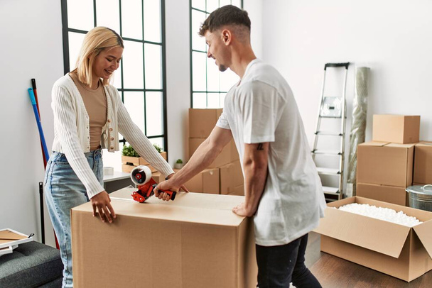 Young caucasian couple smiling happy closing box using packing tape at new home. - Photo, Image