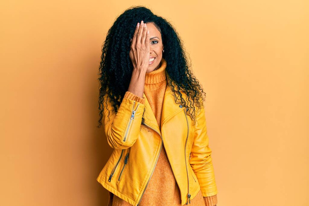 Středního věku africký Američan žena na sobě vlny zimní svetr a kožené sako zakrývající jedno oko s rukou, sebevědomý úsměv na tváři a překvapení emoce.  - Fotografie, Obrázek