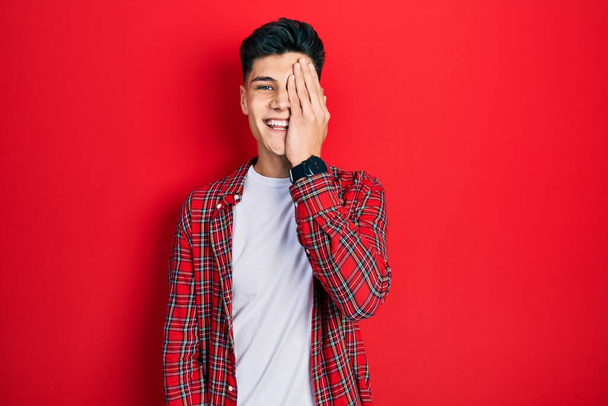 Jeune homme hispanique portant des vêtements décontractés couvrant un oeil avec la main, sourire confiant sur le visage et l'émotion surprise.  - Photo, image