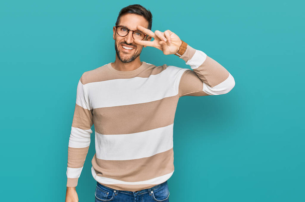 Handsome man with beard wearing casual clothes and glasses doing peace symbol with fingers over face, smiling cheerful showing victory  - Photo, Image