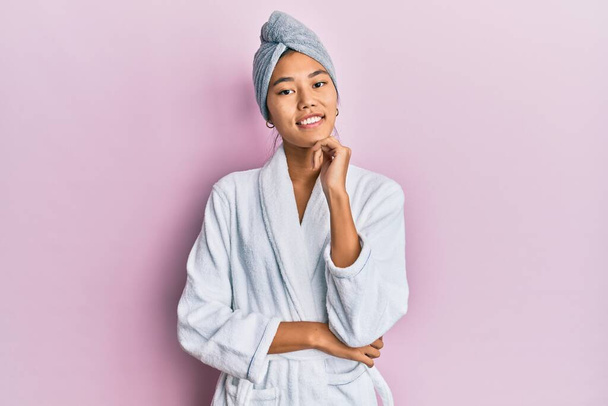 Young chinese woman wearing shower towel cap and bathrobe looking confident at the camera with smile with crossed arms and hand raised on chin. thinking positive.  - Φωτογραφία, εικόνα