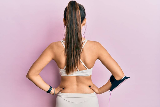 Young hispanic woman wearing gym clothes and using headphones standing backwards looking away with arms on body  - Foto, Imagem