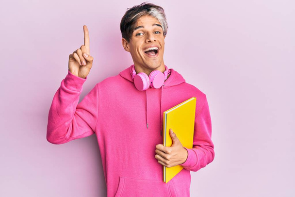 Homem hispânico jovem segurando livro usando fones de ouvido sorrindo com uma ideia ou pergunta apontando dedo com cara feliz, número um  - Foto, Imagem