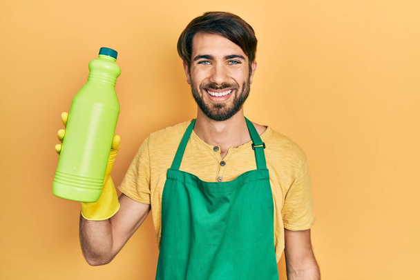 Young hispanic man wearing cleaner apron holding cleaning product looking positive and happy standing and smiling with a confident smile showing teeth  - Фото, изображение