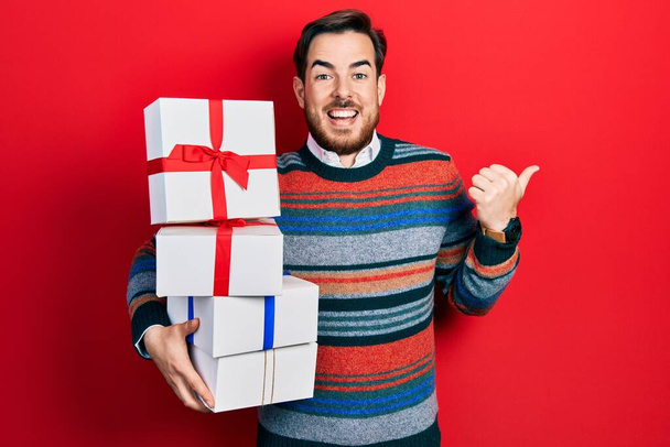 Knappe blanke man met baard met geschenken die duim naar de zijkant wijzen en blij glimlachen met open mond  - Foto, afbeelding