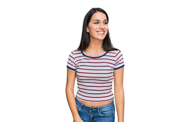 Junges hispanisches Mädchen in lässig gestreiftem T-Shirt, das mit einem Lächeln im Gesicht und natürlichem Ausdruck zur Seite schaut. Lachen selbstbewusst.  - Foto, Bild