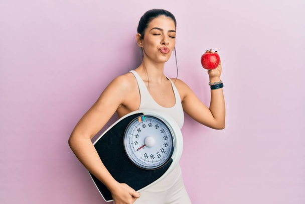 Fiatal spanyol nő visel sportruházat gazdaság súlymérő gép és alma nézi a kamera fúj egy csókot, hogy szép és szexi. szerelem kifejezés.  - Fotó, kép