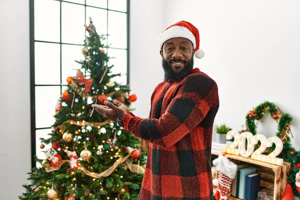 Αφροαμερικάνος φορώντας καπέλο Σάντα Claus στέκεται από το χριστουγεννιάτικο δέντρο δείχνοντας στην άκρη με τα χέρια ανοιχτά παλάμες δείχνει χώρο αντίγραφο, παρουσιάζοντας διαφήμιση χαμογελώντας ενθουσιασμένος χαρούμενος  - Φωτογραφία, εικόνα