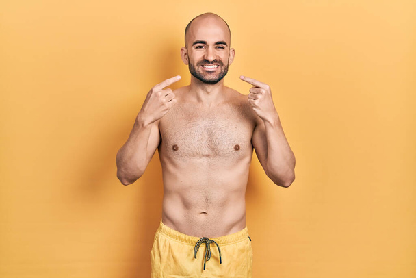Young bald man wearing swimwear smiling cheerful showing and pointing with fingers teeth and mouth. dental health concept.  - Photo, Image