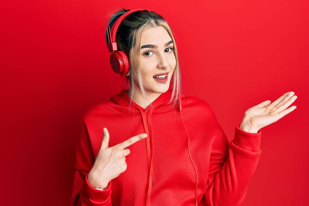 Young modern girl wearing gym clothes and using headphones amazed and smiling to the camera while presenting with hand and pointing with finger.  - Fotó, kép
