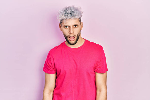 Jeune homme hispanique aux cheveux teints modernes portant un t-shirt rose décontracté au visage choc, à l'air sceptique et sarcastique, surpris avec la bouche ouverte  - Photo, image