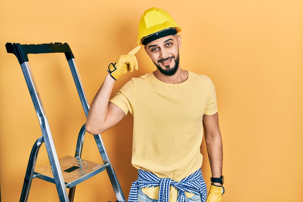 Handsome man with beard by construction stairs wearing hardhat smiling pointing to head with one finger, great idea or thought, good memory  - Photo, Image