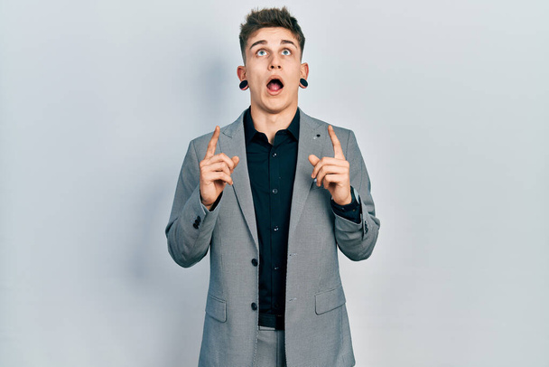 Young caucasian boy with ears dilation wearing business jacket amazed and surprised looking up and pointing with fingers and raised arms.  - Фото, изображение