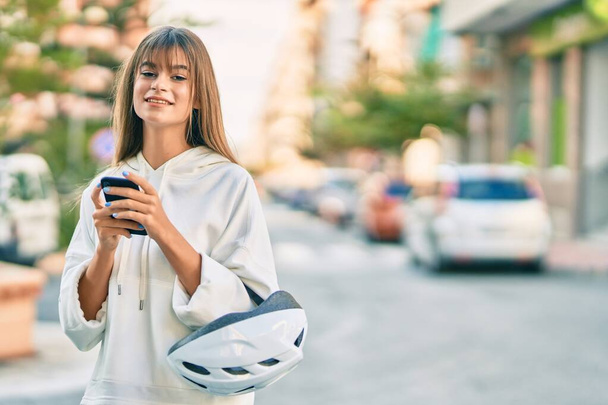 白人スポーティなティーン女の子保持自転車ヘルメットを使用してスマートフォンで街. - 写真・画像