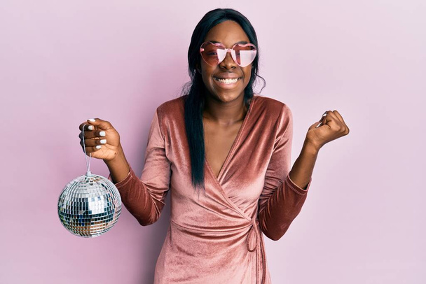 Молодая африканская американка в сексуальном платье для вечеринки, держа диско-мяч, гордо крича, празднуя победу и успех, очень взволнована поднятой рукой  - Фото, изображение
