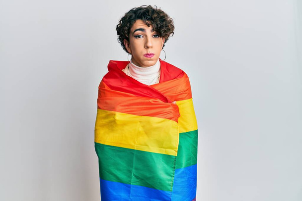 Jovem vestindo mulher compõem envolto em arco-íris lgbtq bandeira soprando bochechas com rosto engraçado. boca inflada com ar, apanhar ar.  - Foto, Imagem