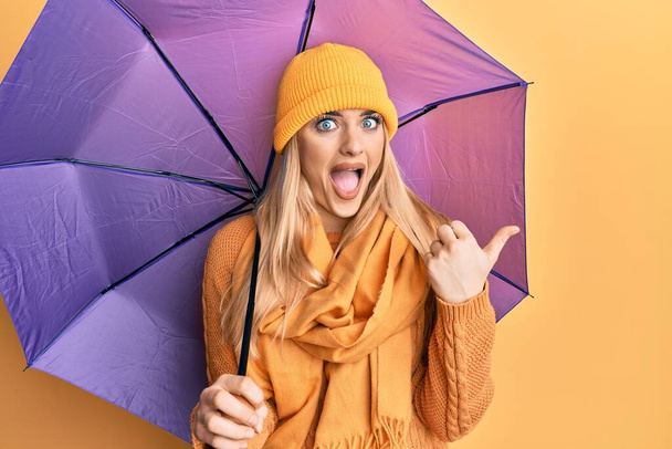 Young caucasian woman holding purple umbrella pointing thumb up to the side smiling happy with open mouth  - Photo, Image