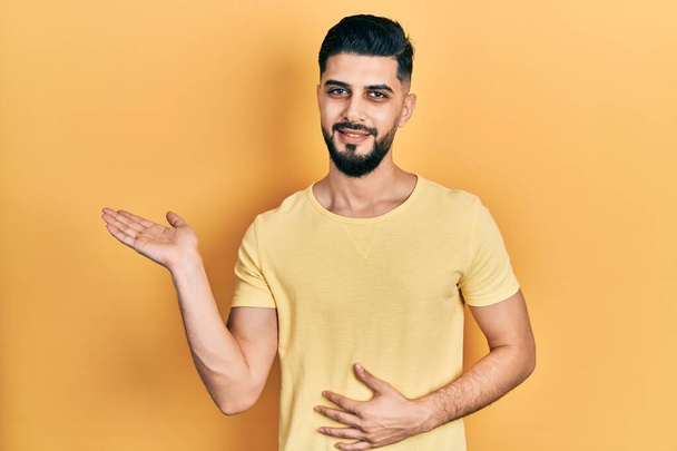 Handsome man with beard wearing casual yellow t shirt smiling cheerful presenting and pointing with palm of hand looking at the camera.  - Фото, изображение