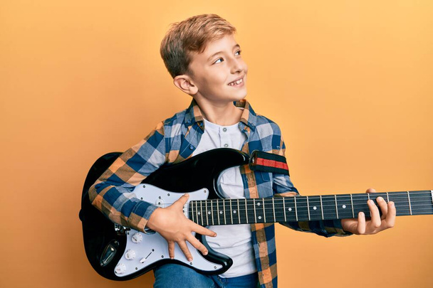 Pequeño niño caucásico tocando la guitarra eléctrica sonriendo mirando hacia un lado y mirando hacia otro pensando.  - Foto, imagen