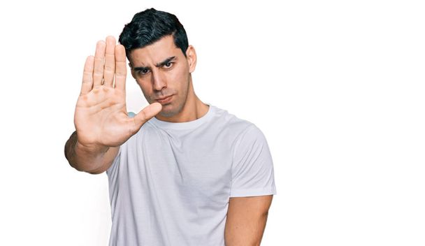 Un bel homme hispanique portant un t-shirt blanc décontracté arrête de chanter avec la paume de la main. expression d'avertissement avec geste négatif et sérieux sur le visage.  - Photo, image