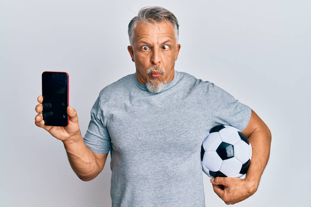 Мужчина средних лет с седыми волосами, держащий футбольный мяч, показывает смартфон, делающий рыбье лицо с ртом и щурящимися глазами, сумасшедший и комичный.  - Фото, изображение