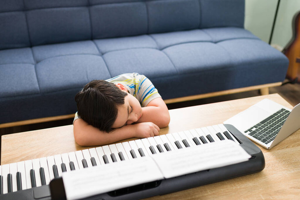 Menino caucasiano entediado dormindo durante suas aulas de música online. Criança cansada adormecendo enquanto ouve seu professor de piano em uma videochamada  - Foto, Imagem