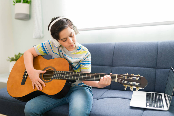 Ταλαντούχο αγόρι ακούει ένα τραγούδι με ακουστικά ενώ παίζει ακουστική κιθάρα και ακολουθεί ένα βίντεο φροντιστήριο στο laptop του - Φωτογραφία, εικόνα