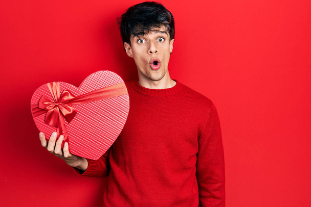 Handsome hipster young man holding valentine gift scared and amazed with open mouth for surprise, disbelief face  - Photo, Image