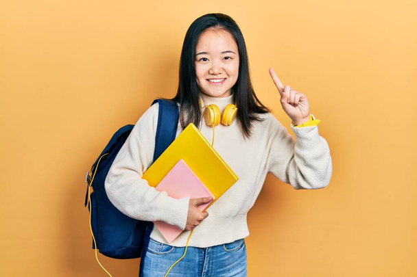 Giovane ragazza cinese tenendo zaino studente e libri puntando il dito con l'idea di successo. Uscito e felice. numero uno.  - Foto, immagini