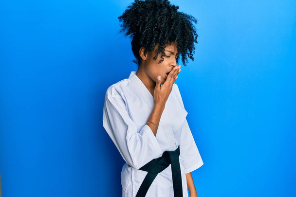 Африканська американка з волоссям афроамериканців, одягнена в карате кімоно і чорний пояс, позіхала від утоми, прикриваючи рот рукою. неспокійний і сонний.  - Фото, зображення