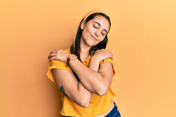 若いヒスパニック系の女性が自分自身幸せと肯定的な抱擁カジュアルな黄色のtシャツを着て、自信を笑顔。自己愛と自己ケア  - 写真・画像