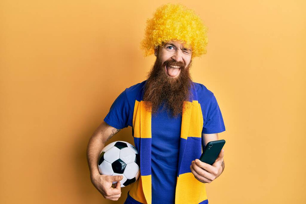 長い髭サッカーのフーリガン応援ゲームを持つ赤毛男セクシーな表情でカメラを見て、陽気で幸せな顔を持つスマートフォンのウインクを保持.  - 写真・画像