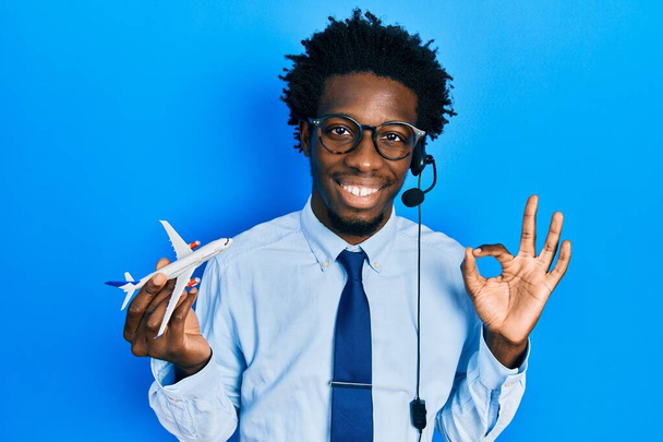 Jonge Afro-Amerikaanse reisagent man houdt vliegtuig doet ok teken met de vingers, glimlachend vriendelijk gebaar uitstekend symbool  - Foto, afbeelding