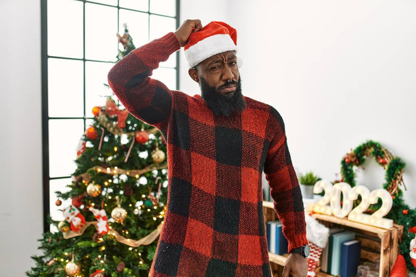 Αφροαμερικάνος που φοράει καπέλο Σάντα Κλάους και στέκεται δίπλα στο χριστουγεννιάτικο δέντρο μπερδεύεται και αναρωτιέται για την ερώτηση. αβέβαιος με αμφιβολία, σκεπτόμενος με το χέρι στο κεφάλι. Σκεπτική έννοια.  - Φωτογραφία, εικόνα