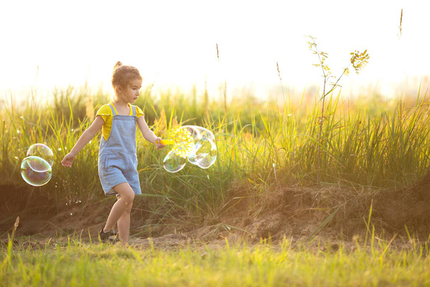 A girl in a denim jumpsuit blows soap bubbles in the summer in a field at sunset. International Children's Day, happy child, outdoor activities. Summer background. Healthy and eco-friendly lifestyle - Fotoğraf, Görsel