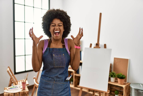 Jeune femme afro-américaine aux cheveux afro au studio d'art célébrant fou et fou de succès avec les bras levés et les yeux fermés hurlant excité. concept gagnant  - Photo, image