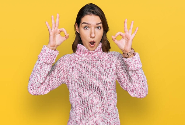 Młoda, kaukaska dziewczyna nosząca wełniany zimowy sweter wyglądająca na zaskoczoną i zszokowaną robiąc ok symbol aprobaty palcami. szalone wyrażenie  - Zdjęcie, obraz