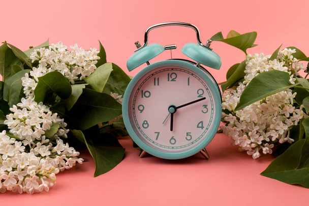 Pembe arka planda izole edilmiş beyaz leylak çiçekli mavi çalar saat. Bahar Zamanı Değişimi, Bahar Çiçekleri ve Alarm Saati.  - Fotoğraf, Görsel