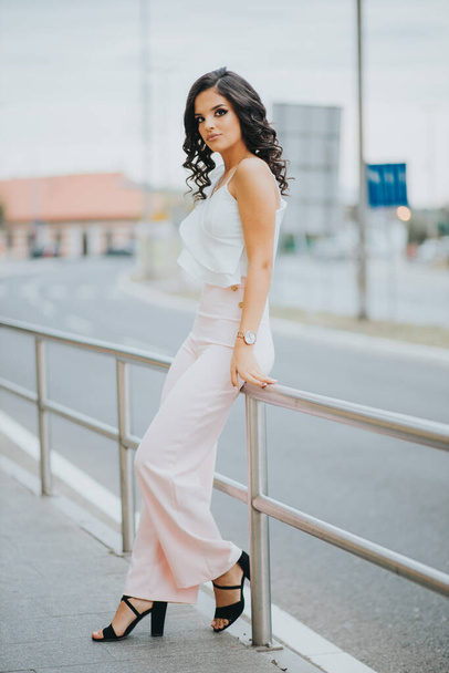 A gorgeous female in an elegant pink and white outfit leaning on an urban street fence - Fotografie, Obrázek