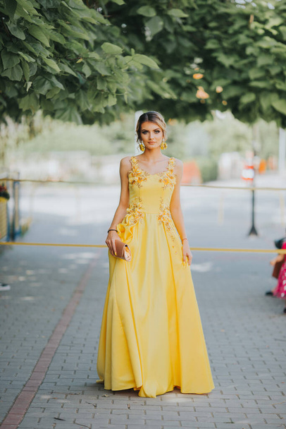 A vertical portrait of a gorgeous Caucasian blonde female holding her elegant yellow dress in street - Photo, image