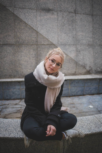 A vertical shot of a blonde girl wearing glasses and a winter coat with a scarf - Foto, Imagen