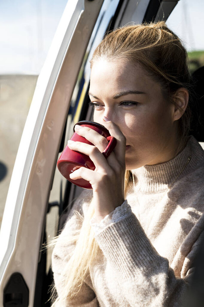 A young female sitting in a car drinking a cup of hot coffee and enjoying the weather - 写真・画像