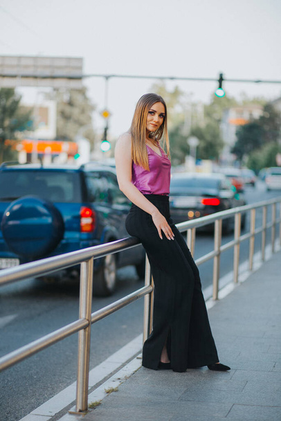 A gorgeous Caucasian female in stylish purple and black outfit leaning on a street fence - Zdjęcie, obraz