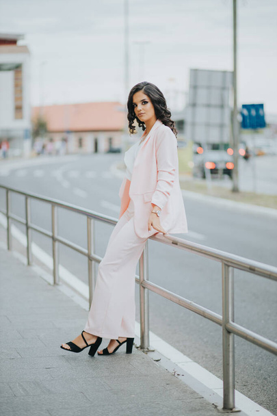 A gorgeous Caucasian female in an elegant pink and white outfit leaning on an urban street fence - Foto, imagen