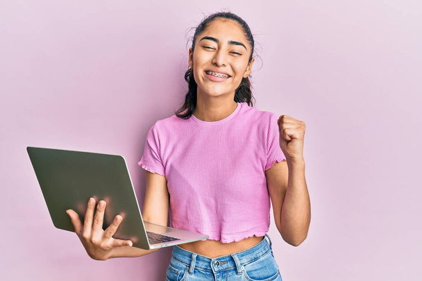 Adolescente hispanique avec des appareils dentaires tenant et utilisant ordinateur portable criant fier, célébrant la victoire et le succès très excité avec le bras levé  - Photo, image