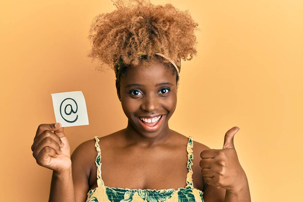 Νεαρή Αφρικανή γυναίκα με αφρο μαλλιά κρατώντας online σύμβολο αλληλογραφίας σε χαρτί χαμογελώντας χαρούμενος και θετικός, αντίχειρας επάνω κάνει εξαιρετική και την έγκριση σημάδι  - Φωτογραφία, εικόνα