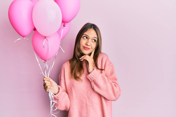 Young brunette woman holding pink balloons serious face thinking about question with hand on chin, thoughtful about confusing idea  - Photo, Image
